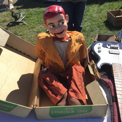 A ventriloquist dummy of Paul Winchell's Jerry Mahoney in its oriignal box sits on a flea market table next to an electric guitar, with green grass in the background.