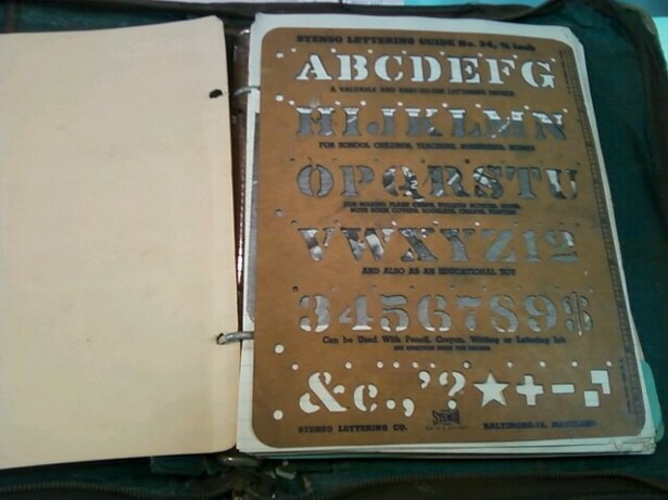 Image description: interior of a scrapbook shows, at left, a blank beige page, and at right, a brown stencil, about standard 8 1/2" by 11" binder size. Stencil contains the alphabet, numbers 1 through 9, and a about a dozen symbols.