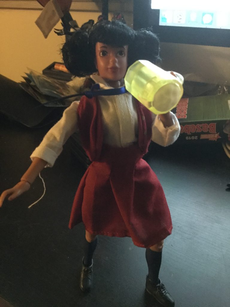 A Mego Tootie action figure from The Facts of Life series, a teenaged looking female doll with black hair and brown skin in a maroon and white private school uniform, drinking a mug of beer.