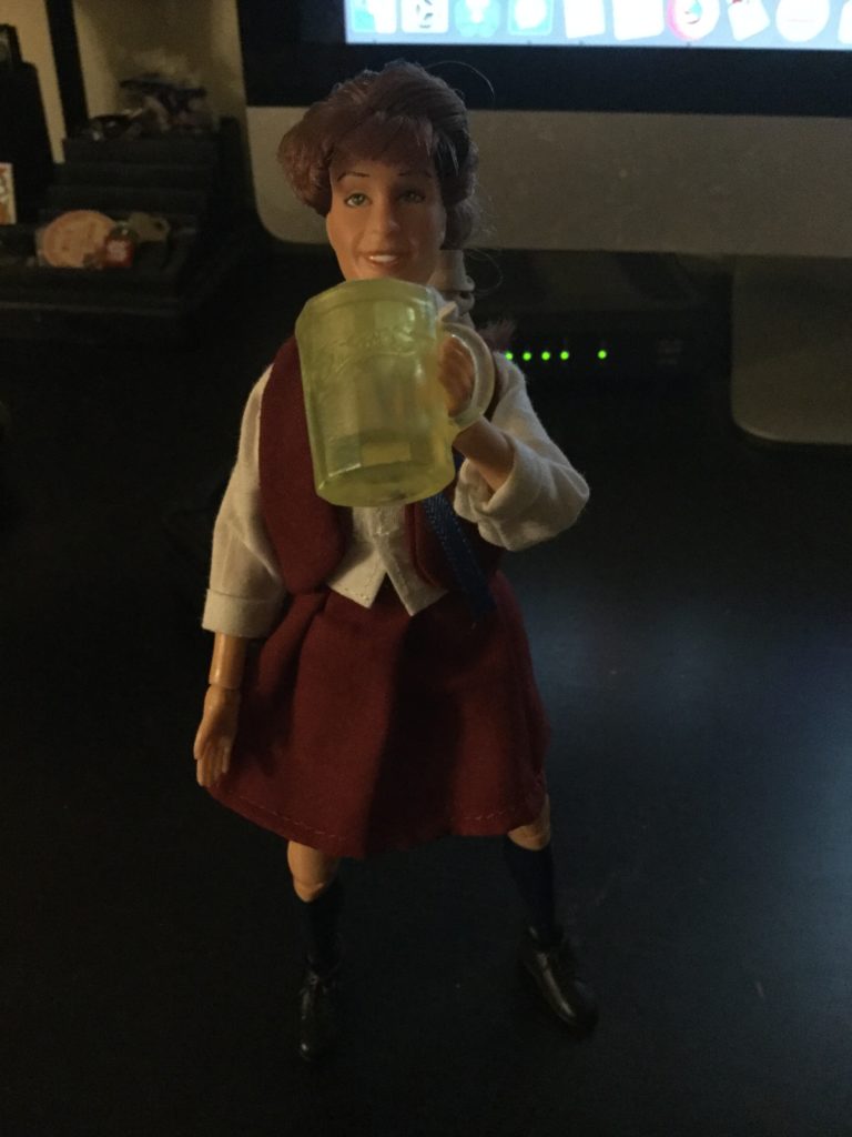 A Mego Jo action figure from The Facts of Life series, a teenaged looking female action figure with white skin and brown hair in a maroon and white private school uniform, drinking a mug of beer.