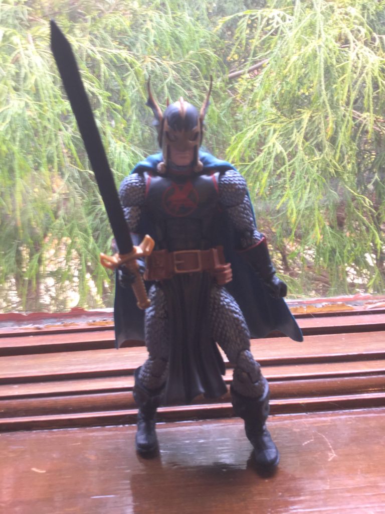 A Marvel Legends Black Knight action figure stands on a wooden window sill, with green foliage in a window behind it. Human figure in a blue tunic with a red insignia and red trim, blue, gold and grey armor, a brown utility belt, and blue boots and gloves, holding a large black sword with a gold hilt.