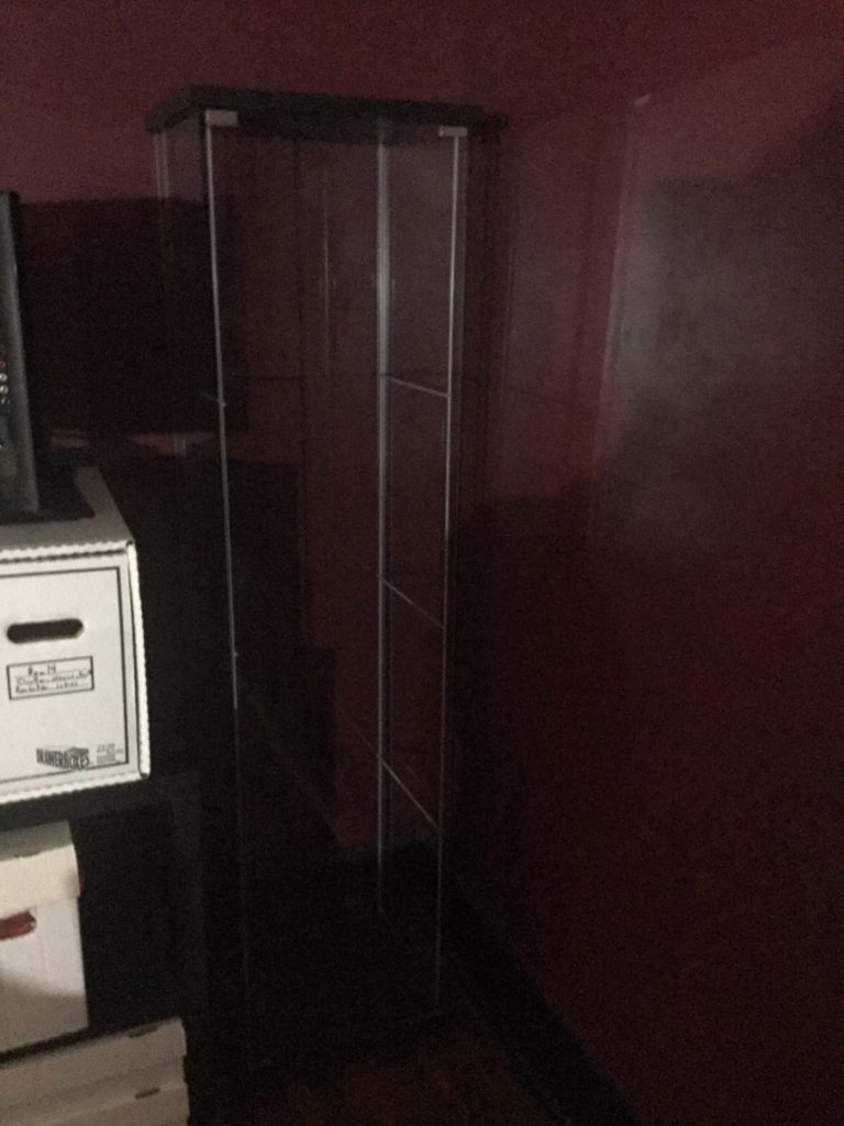 An empty glass display case, center of picture, rests up against maroon walls, with some white boxes on a black shelf to its left.