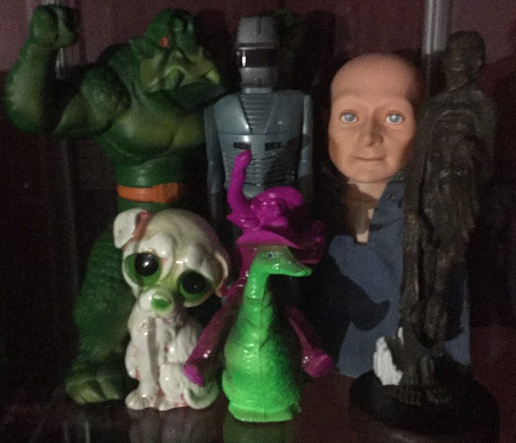 Various toys and sculptures sit on a shelf: Ideal's KRUSHER doll, a green monster with orange trim; ROM The Spaceknight, a grey robot with red trim; Hugo, the Man of a Thousand Faces, a bald puppet with white skin wearing a blue denim shirt; a statue of a vampire version of comic book character The Joker; a green, pink and white ceramic statue of a sad looking dog with large green and black eyes; A green duck wearing a pink cowboy hat, being ridden by a pink cowboy.