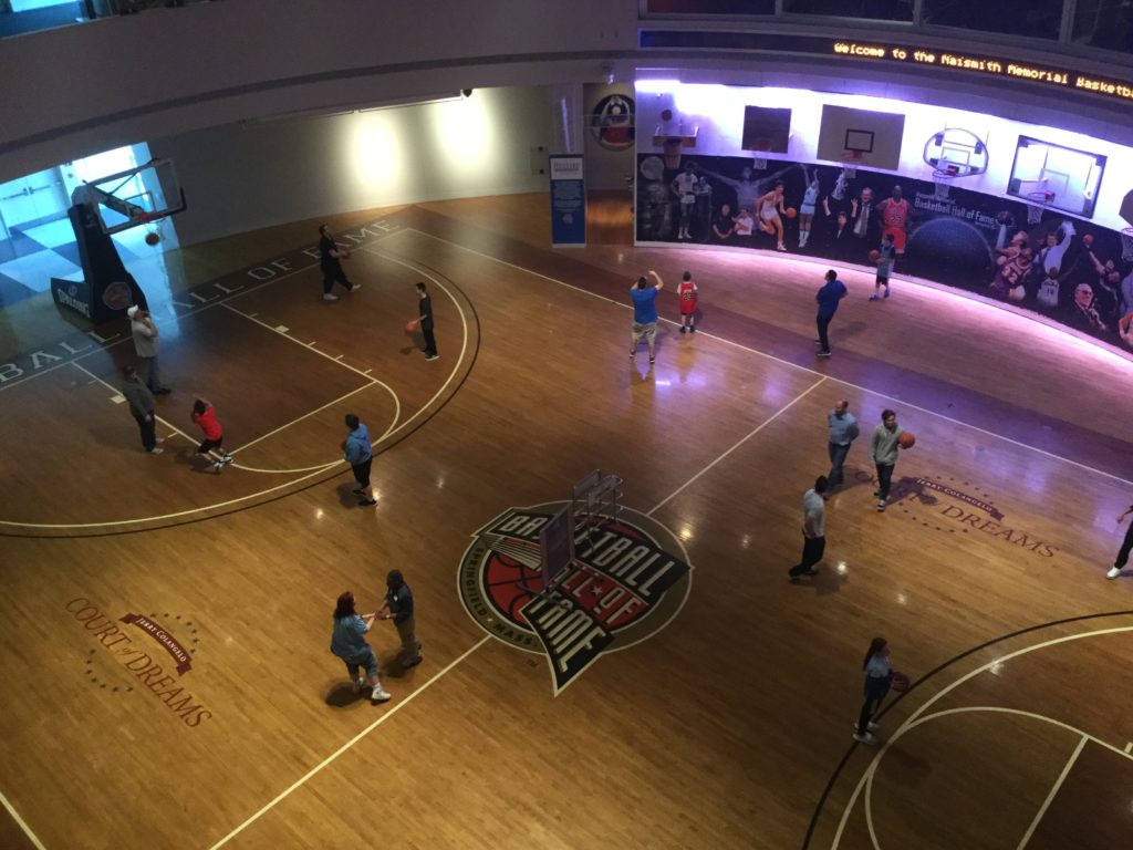 An overhead shot of the basketball courts of the Naismith Memorial Basketball Hall of Fame in Springfield, MA. approximately 15 people play basketball near a number of basketball hoops, in a circular basketball gym with pictures of famous basketball players off in the distance.