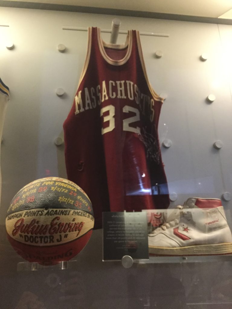 The Julius Erving display case at the Naismith Memorial Basketball Hall of Fame in Springfield, MA. A University of Massachusetts maroon and white basketball jersey, an inscribed red, white and blue basketball, and a white Converse basketball sneaker with red trim are visible in the case, along with a placard that is not legible.