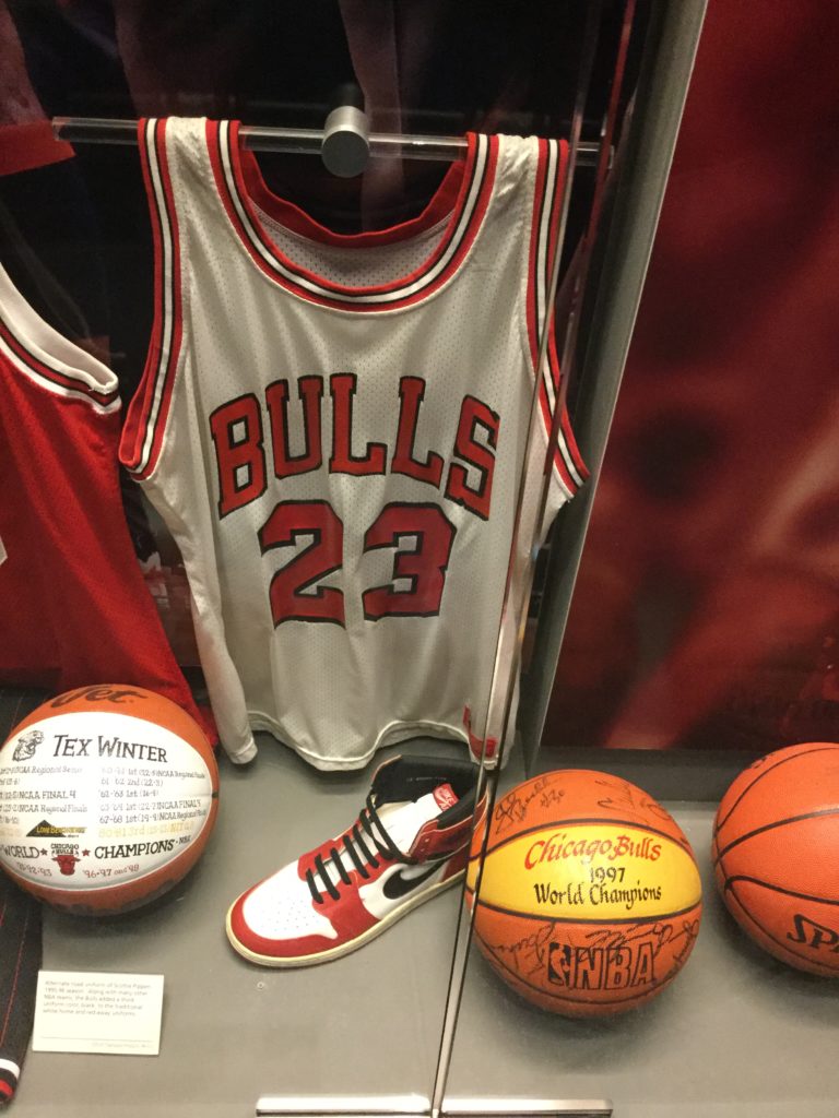 The Michael Jordan display case in the Naismith Memorial Basketball Hall of Fame in Springfield, MA. A Chicago Bulls #23 jersey, white with red trim, once worn by Michael Jordan, a red and white Nike Air Jordan sneaker, and several inscribed basketballs are visible in the case in this picture.