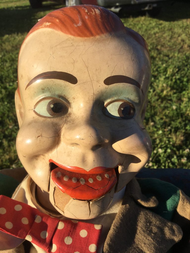 A close-up picture of a decaying Jerry Mahoney ventriloquist dummy's head. White skin, orange hair, brown eyes and eyebrows, red lips, white dots for teeth, red bowtie with white polka dots, brown jacket collar. Paint visibly chipped throughout the face, especially on the jaw.