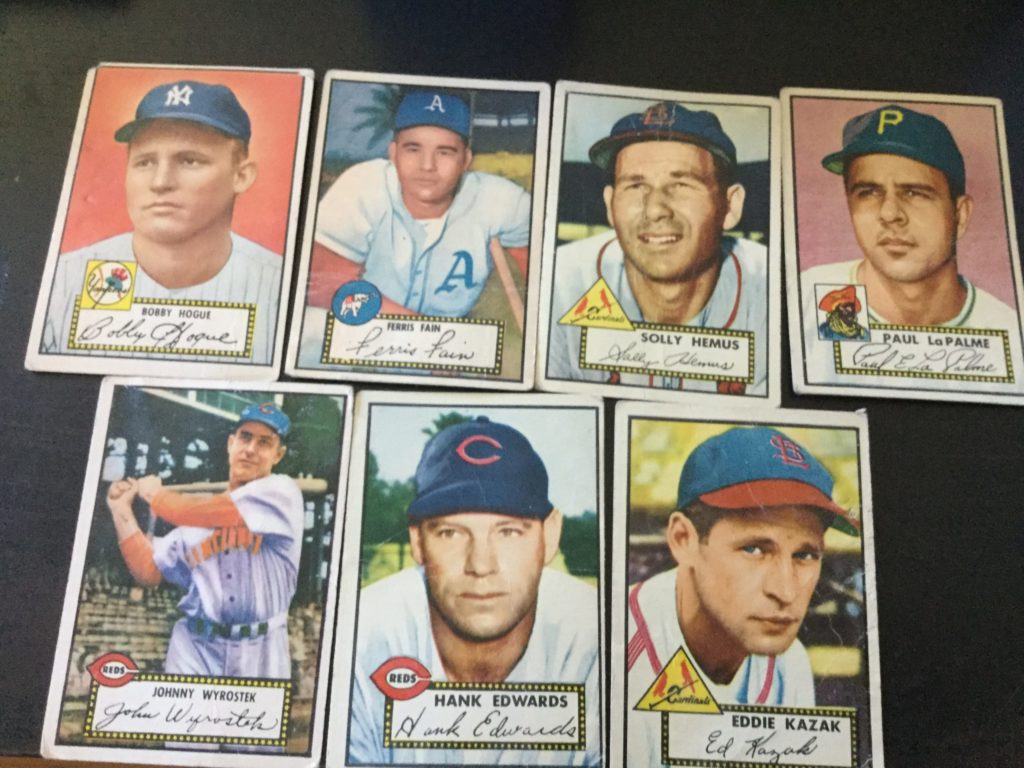 Picture of seven 1952 Topps baseball cards, all featuring men with white skin and brown hair in baseball uniforms, on a black table. Top row: Bobby Hogue (Yankees), Ferris Fain (Athletics), Solly Hemus (Cardinals), Paul LaPalme (Pirates), pictured as painted portraits against various backgrounds. Bottom row: Johnny Wyrostek (Reds) swinging a baseball bat, 3/4 full body shot), Hank Edwards (Reds), and Eddie Kazak (Cardinals), the last two being portrait shots against various backgrounds.