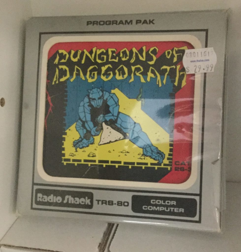 A picture of a boxed Dungeons of Daggorath cartridge for Radio Shack's TRS-80 Color Computer. a blue-gray gargoyle points a sword at the camera in a maze with a yellow floor and bluew walls, with yellow and black Dungeons of Daggorath logo floating above him. Picture is framed by red trim. Box of cartridge is grey, with black trim and lettering around the outside, and white inner window trim near the window the cartridge is displayed in.