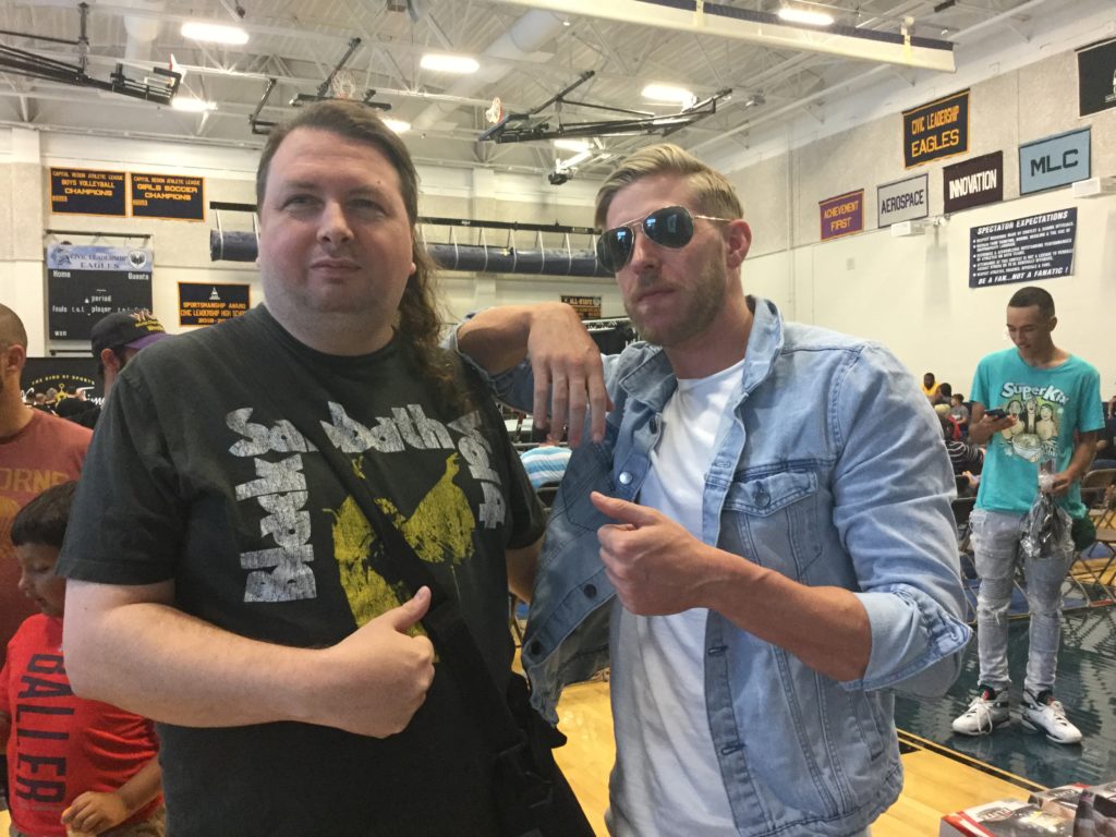 The author (left, a man with brown hair and white skin, wearing a black Black Sabbath Volume IV t-shirt) and pro wrestler Orange Cassidy (right, a man with reddish blonde hair and white skin, wearing aviator sunglasses, a blue jean jacket, and a white t-shirt) give very mild thumbs ups to the camera at a Blitzkrieg Pro/Big Time Wrestling event, August 24th, 2019. Photo Credit: Bryce Remsburg