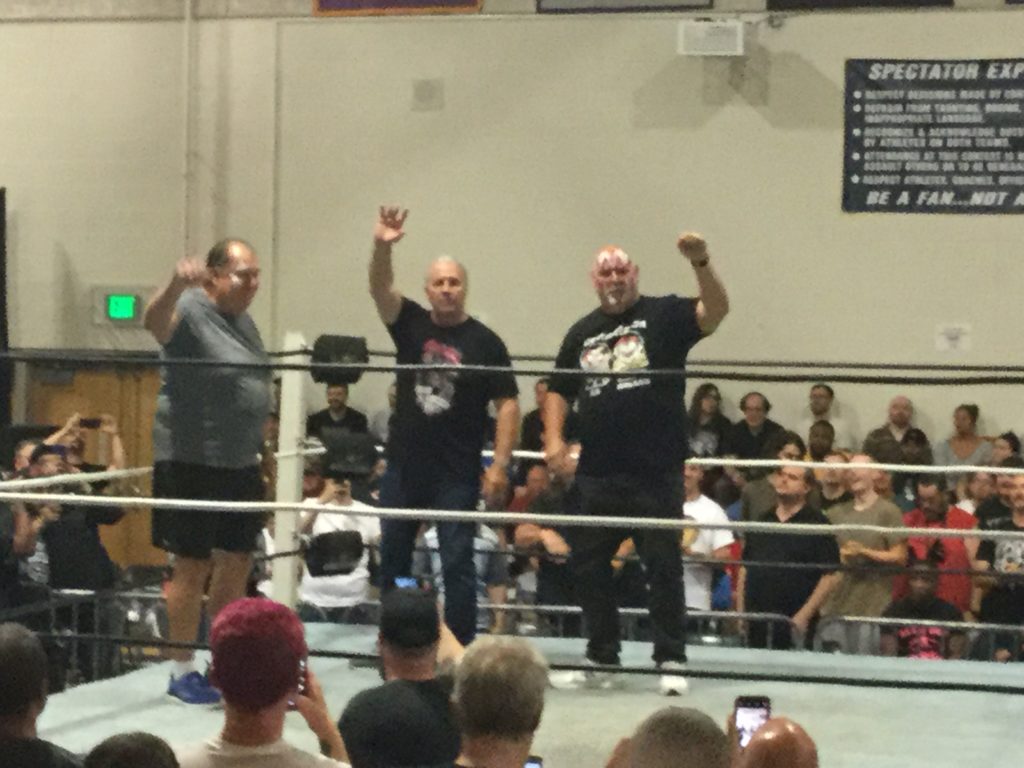 Demolition Ax (Bill Eadie, left, a man with black hair and white skin wearing a grey t-shirt, shorts and blue sneakers), Bret "The Hitman" Hart (center, a man with grey hair and white skin, wearing a black t-shirt and black jeans) and and Demolition Smash (Barry Darsow, right, a bald man with white skin, wearing a black t-shirt, black jeans and white sneakers) stand in a wrestling ring and wave to the crowd at a Blitzkrieg Pro/Big Time Wrestling event at CREC Civic Leadership High School in Enfield, CT on August 24th, 2019.