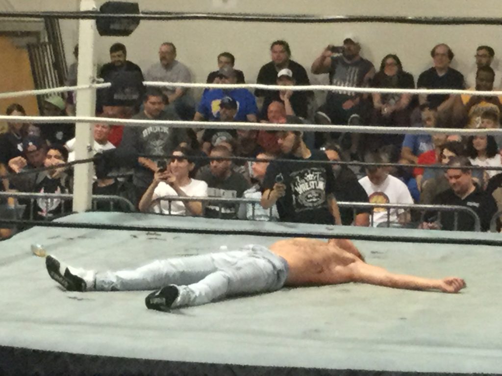 Professional wrestler Orange Cassidy (center, a shirtless man with white skin, wearing blue jeans and white sneakers with black soles) lies motionless in a wrestling ring as a crowd looks on at a Blitzkrieg Pro/Big Time Wrestling event, August 24th, 2019.