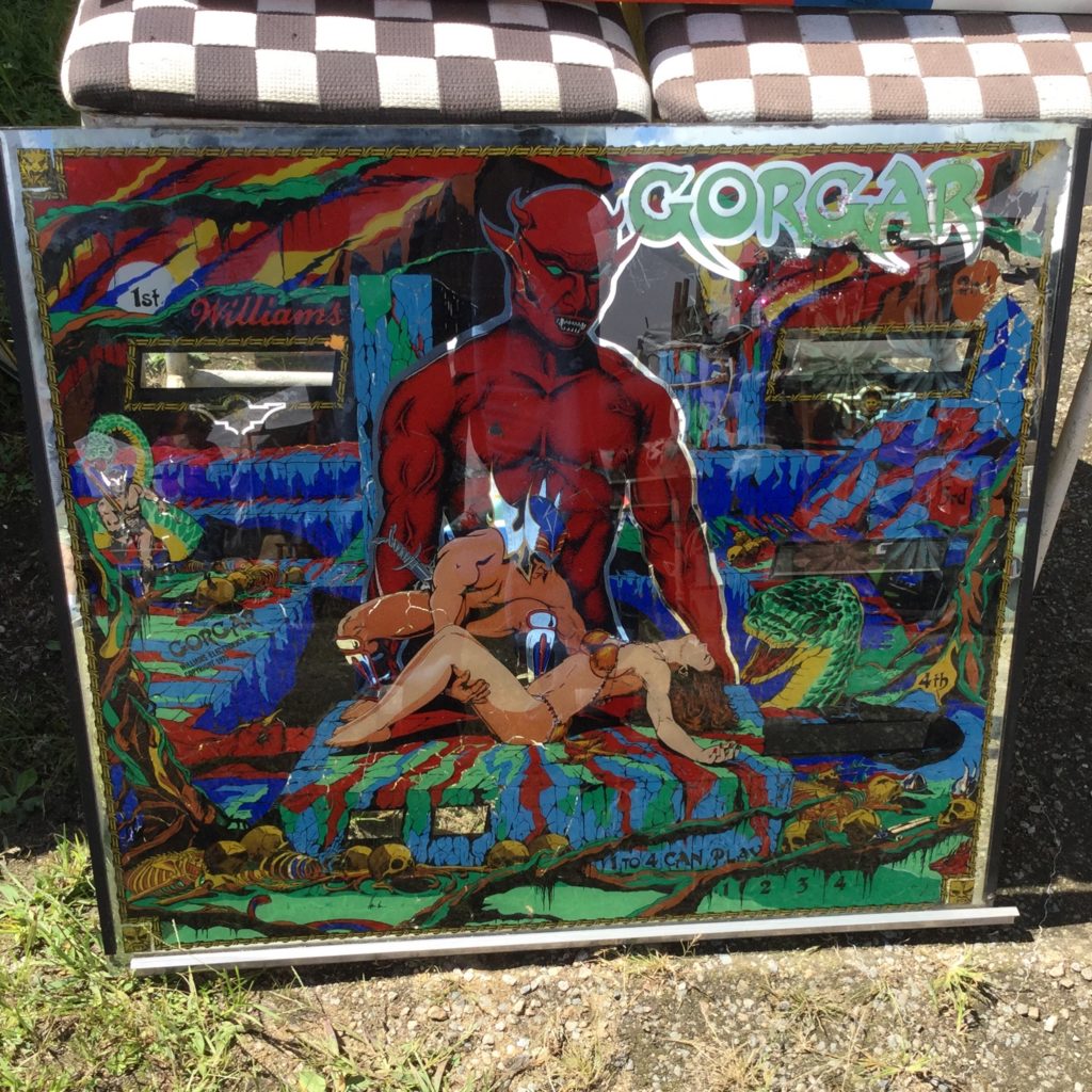 Glass back panel for a 1979 Gorgar pinball machine by Williams. A red devil stands behind a scantily-clad male barbarian with white skin, wearing a blue helmet, who is trying to rescue a scantily clad woman with white skin and brown hair from a bloody altar. "GORGAR" is written in teal lettering with silver trim at the top right.