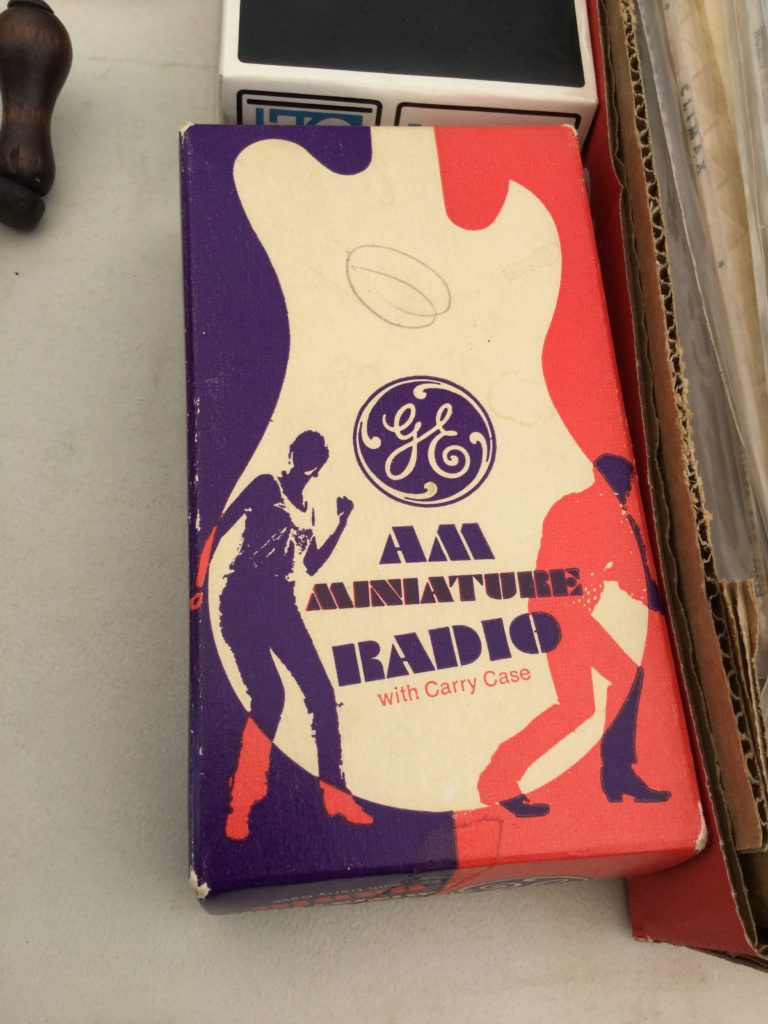 A box for a GE AM Miniature Radio with Carry Case from the late 1960s. On it, there are silhouettes of a man and woman dancing, and the body of an electric guitar, both inset on a background that's purple on the left side and orange on the right. The parts of the man and woman that show up against the backgrounds instead of the guitar are the opposite color of the main background (orange for left, purple for right).