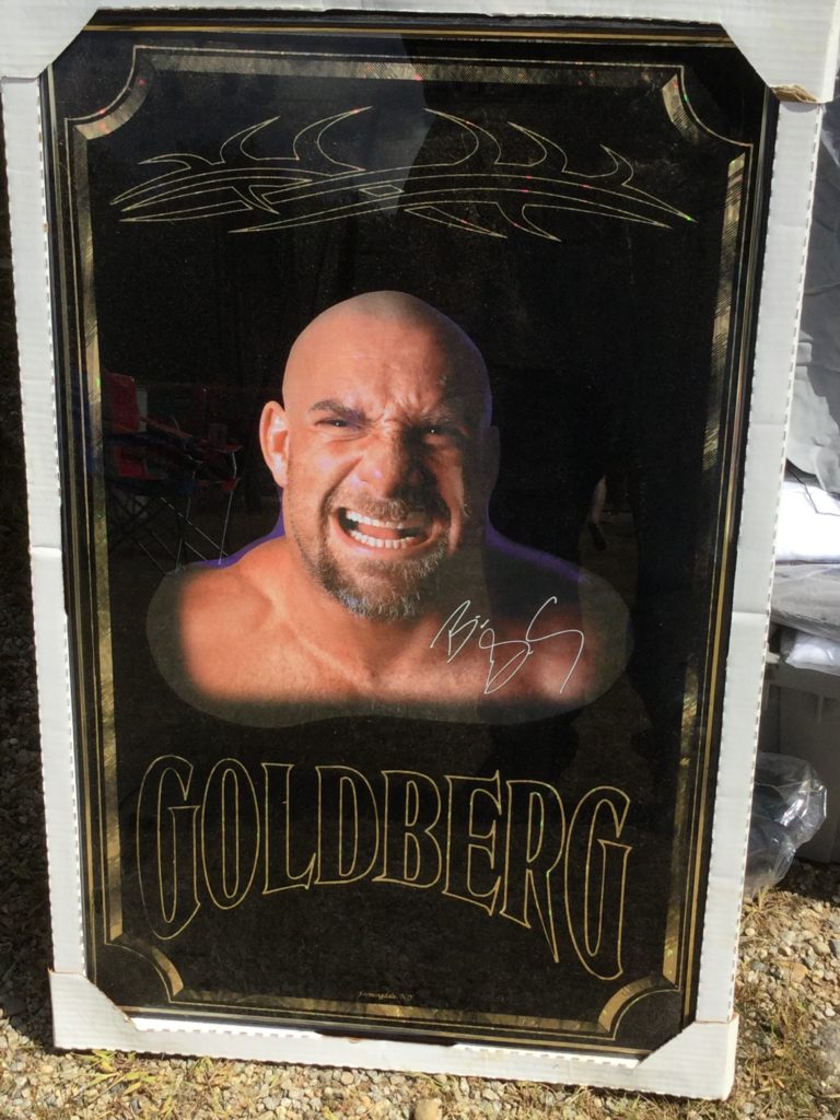 A framed picture of professional wrestler Bill Goldberg, a man with white skin, no hair and a greying brown beard, photographed from the shoulders up. The photo is set against a black background with gold trim. Above his head is a facsimile of the tribal tattoo Goldberg has on his bicep, and below his shoulders, "GOLDBERG" is written in large black letters with gold trim.