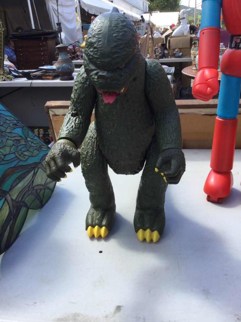 A 1970s Mattel Godzilla toy (a green action figure-type toy standing about 18" high with yellow claws, teeth and eyes, and a red strip of fire in its mouth that resembles a tongue) stands on a table, with a blue and green glass Tiffany lamp to its left, and a Mattel Shogun Warriors Dragun toy (also a large action figure, but a blue, red and black robot) to its right. More tables of antiques are visible in the background.