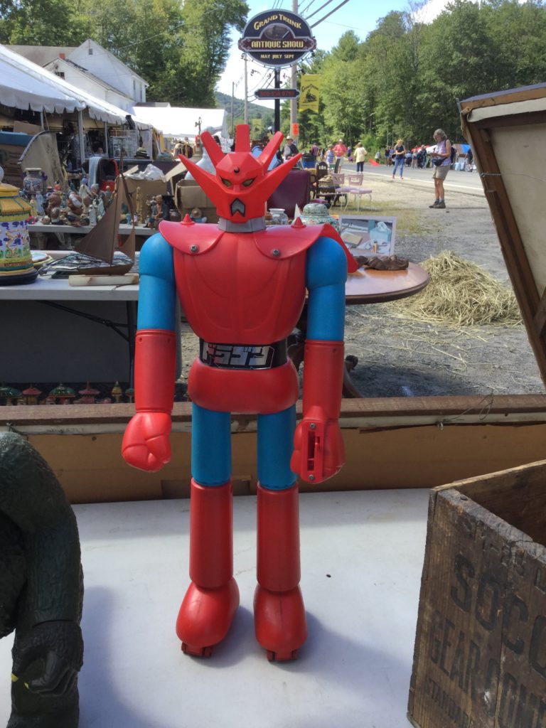 A 1970s Mattel Shogun Warriors Dragun toy ( a red, blue, black and silver robot action figure toy standing about 18" high) stands on a table, with a wooden box to its left, and a Mattel Godzilla toy to its right. More tables of antiques are visible in the background.