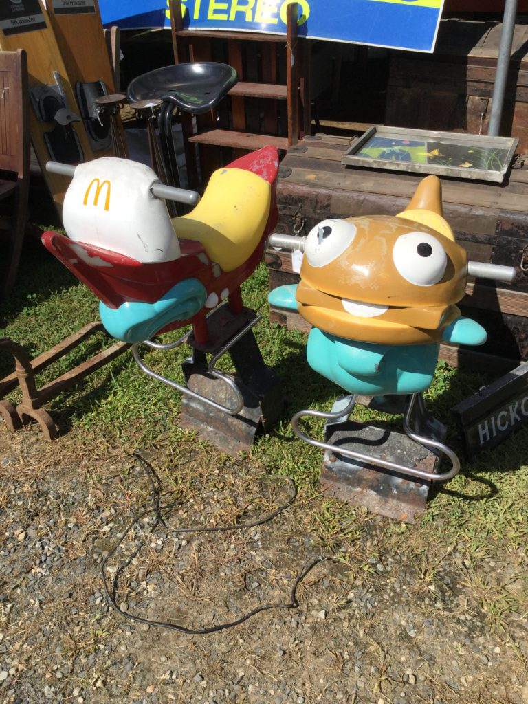 Two small, older McDonald's playground rocking horse-ish rides sit on green grass, with a patch of brown dirt and gravel in front of it at Brimfield Antiques Market, with other antiques in the background. At left, a red white and yellow boat with a McDonald's logo on the mast, silver handles, and a teal simulated sliver of ocean water at the bottom. At right, a similar ride, but the ride is an anthropomorphic Filet-O-Fish sandwich (a brown roll, brown fish filet, white tartar sauce to simulate the appearance of a tongue, wide white eyes with black pupils, and teal fins) instead of a boat.