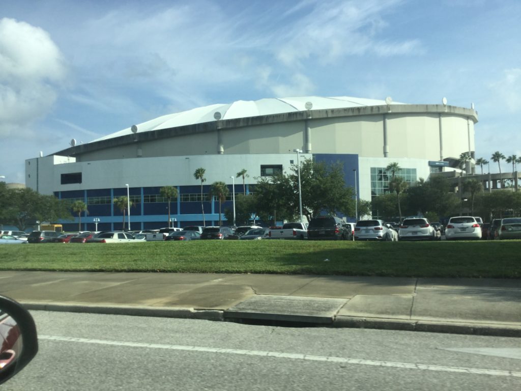 Exterior shot of Tropicana Field in St. Petersburg, Florida. Tropicana Field is an off-white domed baseball stadium with an angled roof that resembles an orange juicer. Cars are parked in front of the stadium, and trees can be seen just past the cars, off in the distance.
