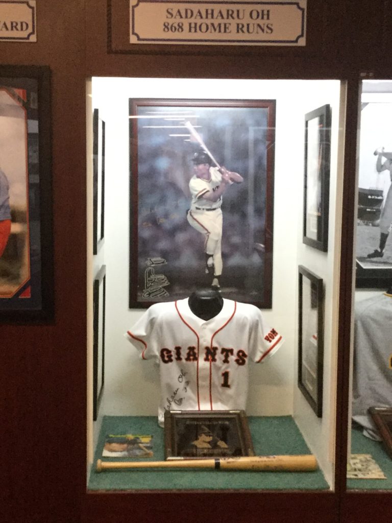 Sadaharu Oh's case in the Hitters Hall of Fame section of the Ted Williams Museum, located at Tropicana Field in St. Petersburg, Florida. At center, a large picture of Sadaharu Oh batting is visible. Just below it, a signed Sadaharu Oh Yomiuri Giants jersey is visible, as well as a magazine, a plaque, and a baseball bat.