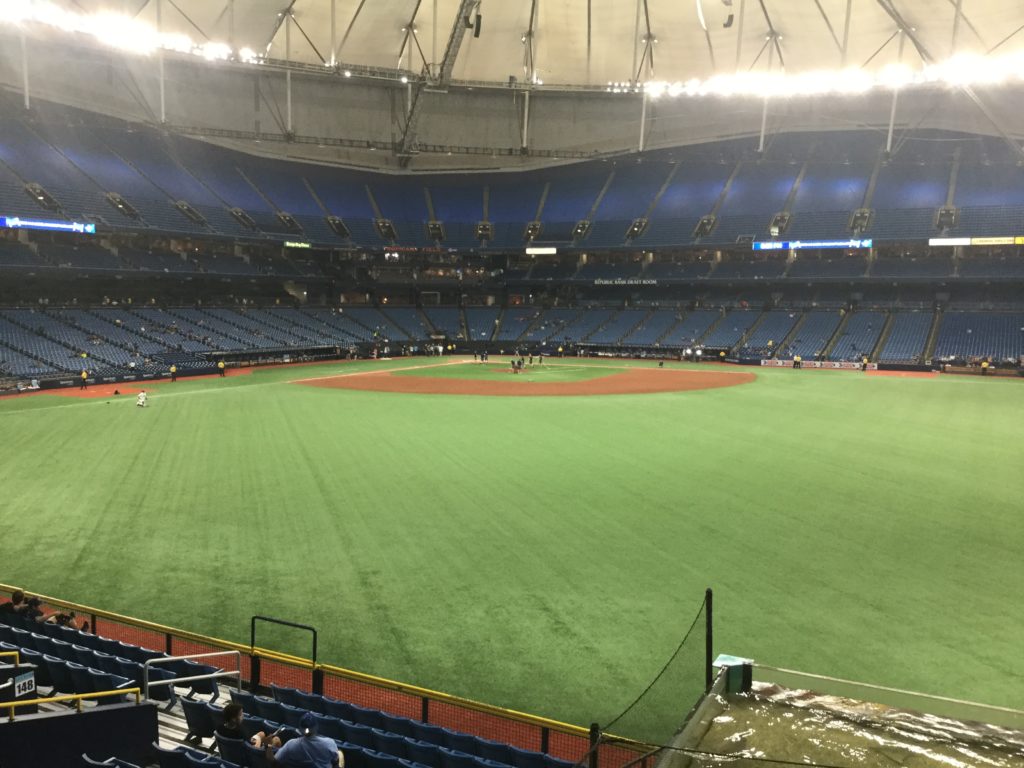 Interior shot of Tropicana Field, taken from right-center field. Green artificial turf and brown infield dirt can be seen, and blue seating is visible on the other side of the field, as well as an off-white roof.