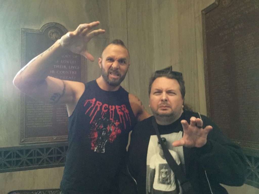 Professional wrestler Lance Archer (left), a tall man with white skin, several arm tattoos, a dark brown beard, a hint of dark brown hair with a red braid weaved into it, and a black t-shirt that says "ARCHER" across the top of it in bloody looking red letters, raises his right hand above his head in a claw position and makes a mean looking face, next to the author of this piece (right), a man with white skin, brown hair with black sunglasses on top of it, a brown goatee, wearing a black hooded sweatshirt and a black t-shirt with a black and white picture of a man on it that's obscured by a black bag strap. The man is also making a mean face, and is making a claw gesture with his left hand. Bronze laques are visible on the marble walls behind the two men.