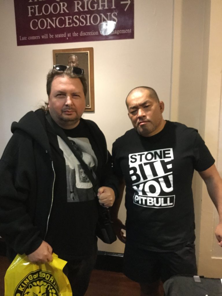 At left, the author, a man with white skin and brown hair (with sunglasses in it), wearing black clothing, holding a yellow New Japan Pro-Wrestling shopping bag. At right, New Japan Pro-Wrestling's Tomohiro Ishii, a stern-looking Japanese man with a bald head, wearing black shorts and a black t-shirt that says "STONE PITBULL BITE YOU" on it.