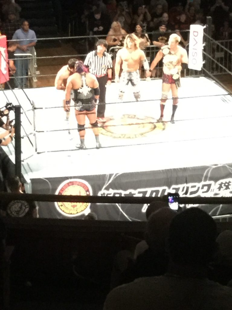 In a wrestling ring, New Japan Pro-Wrestling wrestlers Kota Ibushi (far left, a shirtless Japanese man with short, dark brown hair) and EVIL (just below Kota Ibushi, a Japanese man in black and white singlet with purple ponytail) stand head to head at left, with referee "Red Shoes" Unno (a Japanese man with black hair in black and white referee uniform, black pants, and red shoes) and wrestlers Hiroshi Tanahashi (a Japanese man with long bleached blonde to brown hair, in black and white spandex pants) and Kazuchika Okada to their right (a Japanese man with short bleached blonde hair, in red tights and boots, holding the IWGP Heavyweight Championship belt over his right shoulder).