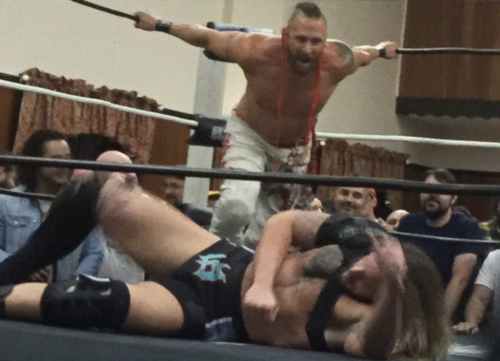 Professional wrestler Lance Archer, a shirtless man with white skin and a red "murderhawk" hairstyle, stands at the back end of a wrestling ring and prepares to attack fellow pro wrestler Josh Briggs, a man with white skin and brown hair in black wrestling trunks and various knee and shoulder braces, laying prone at the front of the wrestling ring, at a Beyond Wrestling show in Worcester, MA.