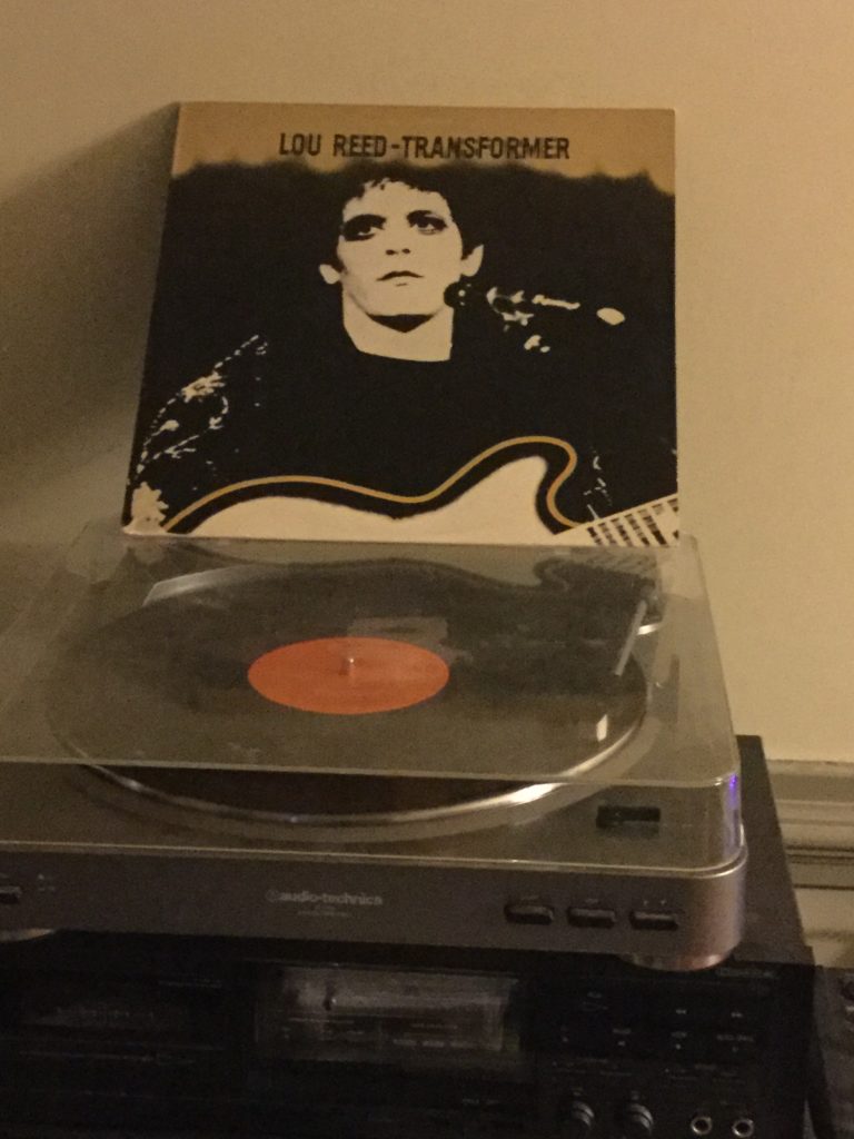 A copy of Lou Reed's "Transformer" album plays on a turntable, with the album cover (featuring a black and white picture of Lou Reed, wearing makeup, and holding a guitar while standing at a microphone) standing on top of the turntable cover.