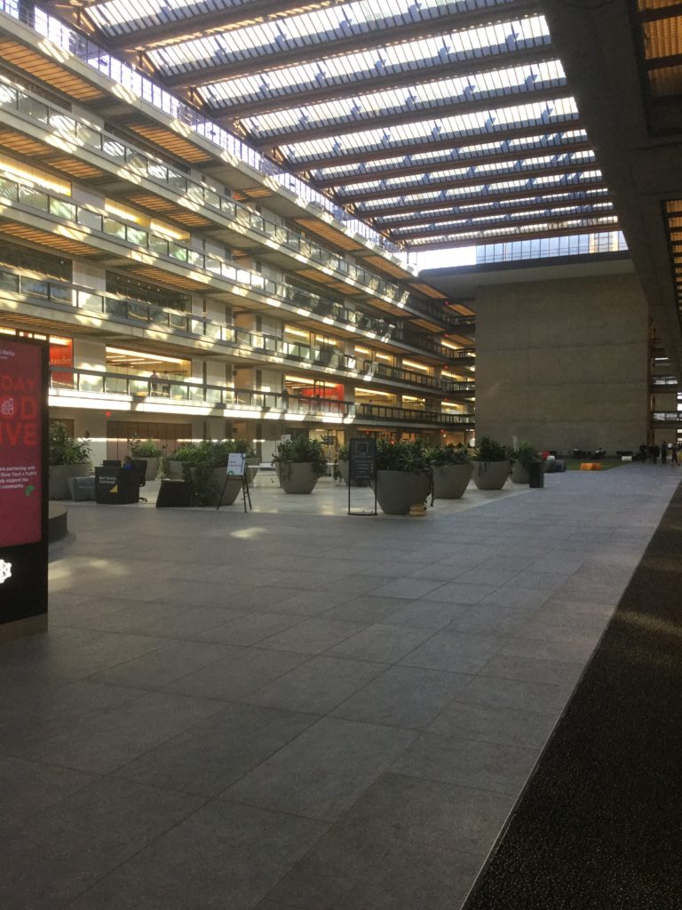 One corridor of Bell Works in Holmdel, NJ. The ceiling is made of glass panels, allowing light into the space. Along the side of the corridor, offices line the space for 6 floors. Planters are visible in the distance on the ground floor, near sitting areas.