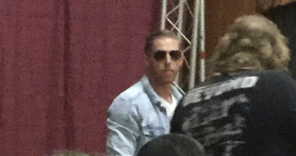 Professional wrestler Orange Cassidy, a man with white skin, red hair, and brown aviator sunglasses, wearing a light blue jean jacket and a white t-shirt, looks directly at the camera from a distance in front of a maroon curtain at a Beyond Wrestling show in Worcester, MA, as a photographer to his right tries to get a picture of him.