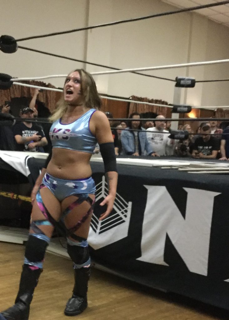 Professional wrestler Kris Statlander, a woman with white skin and brown hair, wearing powder blue wrestling gear with purple trim and "KS" written on her tank top, yells for the crowd at a Beyond Wrestling show in Worcester, MA.