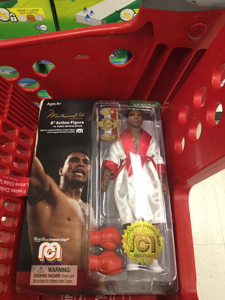 A packaged Mego 8" action figure of boxer Muhammad Ali, a man with brown skin and black hair, wearing a white and red robe, with red boxing gloves and red, white, blue and gold championship belt, rests in a red shopping cart. A picture of Muhammad Ali, right arm raised, is on the card of the package, to the left of the figure.