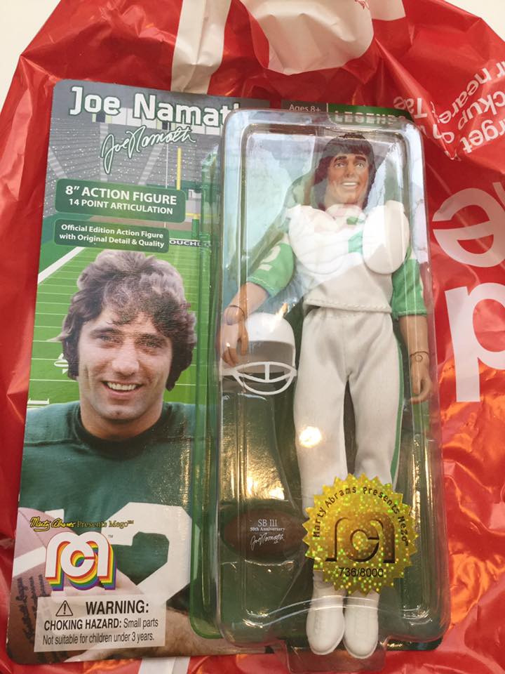 A Mego Joe Namath 8" action figure in its original package rests on top of a red and white bag. Football player Joe Namath, a smiling white man with brown hair, in a white uniform with green trim, with helmet and football. On the package's card, to the left of the figure, there is a picture of Joe Namath in a green uniform with white trim, holding a football.