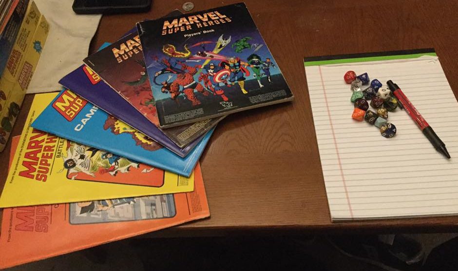 A selection of TSR Marvel Super-Heroes role-playing game handbooks, fanned out to the left, and a white legal pad with a pen and a large array of 10-sided dice to the right, on a brown table.