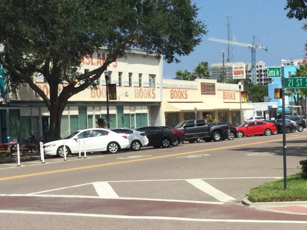 Photo of Haslam's Book Store, St. Petersburg, Florida, taken from across Central Avenue South at 21st Street. A beige building with brick red painted letters that say "HASLAM'S" at the top level (obscured by a tree), and "BOOKS" near the top of the ground level in five different spots. Green curtains cover the windows of the building. A number of cars are parked in front of Haslam's, and the Central Av. S. and 21 St. S. signs are visible across the street.