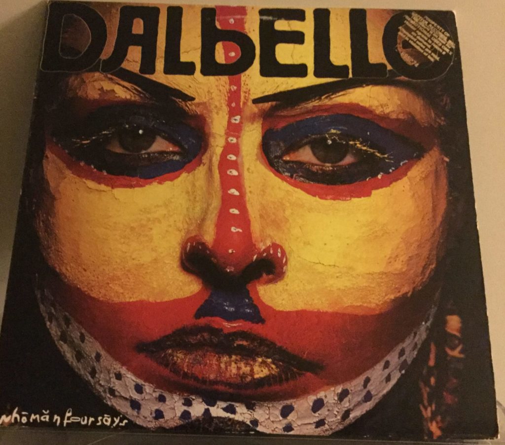 The cover to Dalbello's "whomanfoursays" album. On the cover is a close-up of Lisa Dalbello's face, in yellow, red, blue, black and white tribal warpaint. the name "Dalbello" appears in black faux Cyrillic lettering at the top of the cover, and "whomanfoursays" appears in the bottom left in white scrawled lettering.