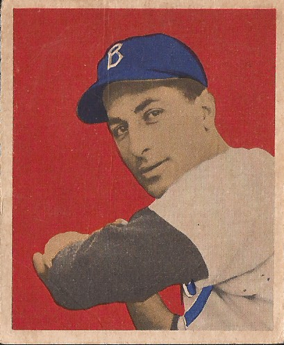 A 1949 Bowman Carl Furillo baseball card. Carl Furillo, a man with white skin and dark hair, wears a white Brooklyn Dodgers uniform with blue trim and a blue baseball cap with the letter B on the front of it, as well as a dark-colored undershirt. Carl's swinging a baseball bat, though only the hilt of the bat is visible. The card is only in partial color, as it was printed in greyscale with the blue highlights on the uniform, against a red background with beige trim.