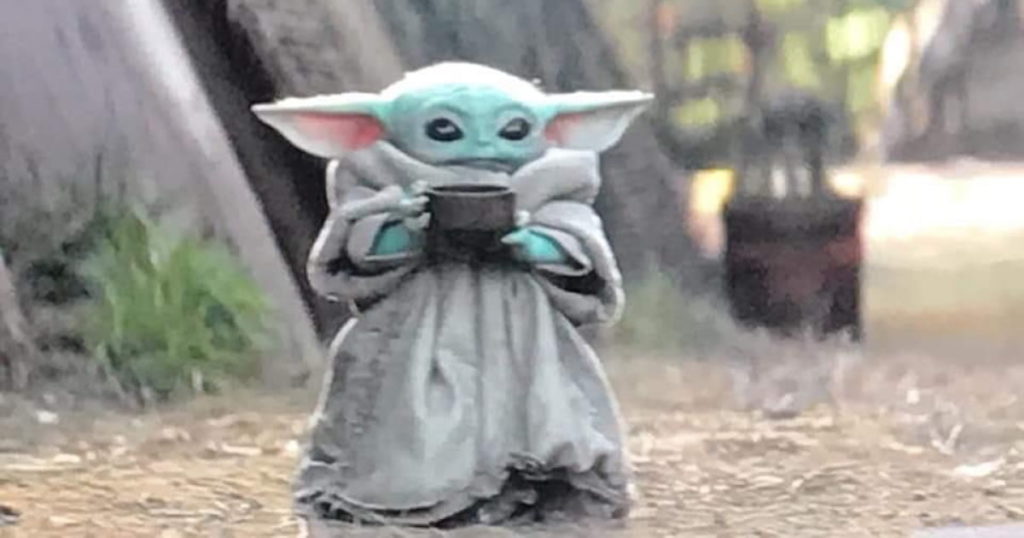 The Child from the television series The Mandalorian, a small green alien with long ears and wide black eyes, wearing a beige robe, sips a mug of soup.